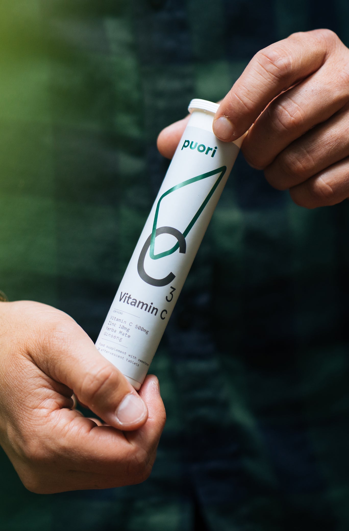 Close-up of two hands holding a Puori C3 Vitamin C effervescent tube against a dark green background.